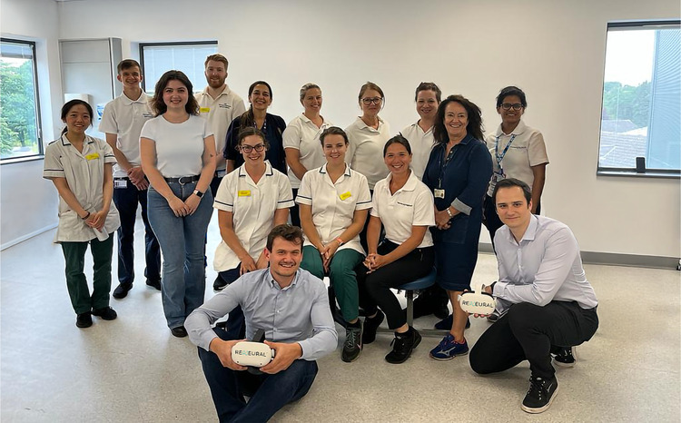 Group photo of the Reneural team and clinical partners in a bright clinical setting. Two people at the front are kneeling and holding VR headsets labelled “RENEURAL,” while others stand smiling behind them, including physiotherapists and staff in uniforms and ID badges.