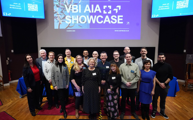 A group of around 20 people stand smiling for a photo on a wooden floor in front of a large screen displaying the text “VBI AIA Showcase” and the date “19 March 2025” at the Bayes Centre. The background screen is purple with a blue abstract design, and promotional QR codes are visible in the top corners. The group appears to be a mix of men and women of varying ages, dressed in smart-casual attire, suggesting a professional event or presentation.